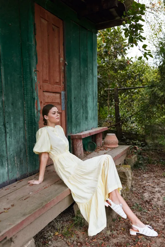 Woman in a pale yellow dress sits on a wooden porch beside a green house in a calm garden setting, creating a serene outdoor fashion portrait