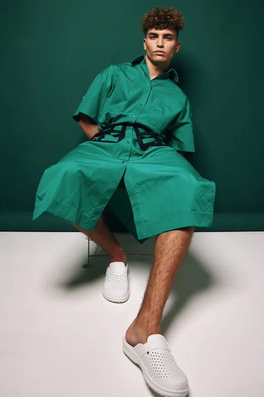 Man wearing oversized green shirt dress and white slip on shoes sitting on chair against green background it fashion style concept casual youth amateur model pose.