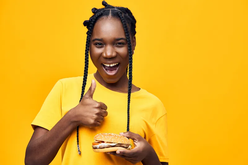 A joyful young Black woman in a bright yellow shirt giving a thumbs up while holding a delicious burger against a vibrant yellow background
