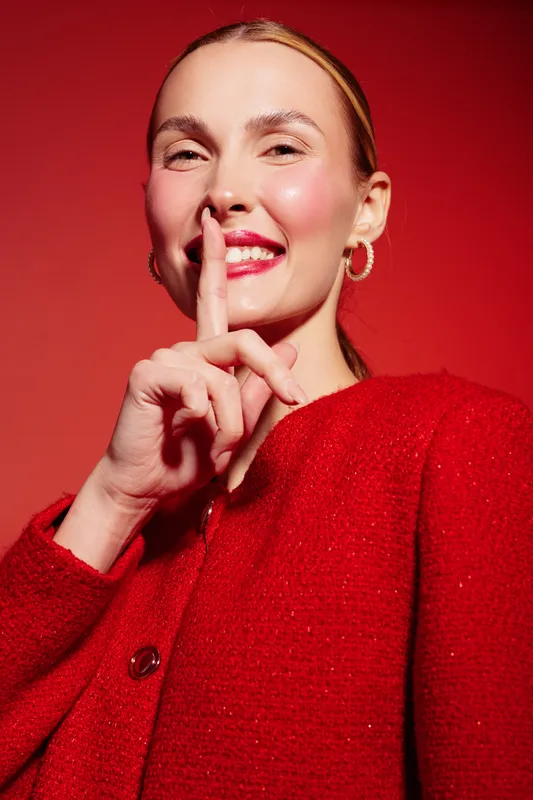 Smiling woman in red sweater makes silence gesture with finger on lips. Close-up portrait on red background. Concept of secrecy, quiet, communication, and positive emotion.