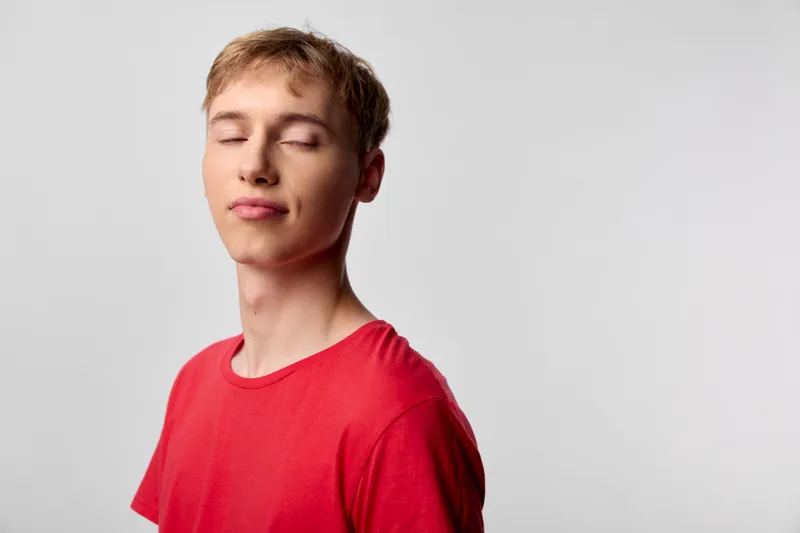 Close up of a young man with eyes closed and a soft smile, wearing a red shirt, in a clean studio, conveying calm, mindfulness and relaxed mood, and serene confidence.