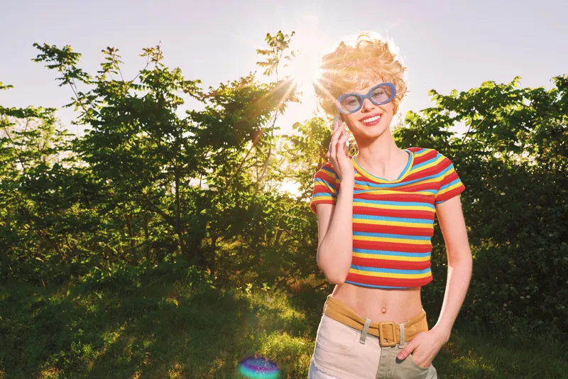 Woman lifestyle portrait in sunny outdoor setting with retro film color, striped crop top, sunglasses, and natural light, conveying cheerful mood and relaxed casual vibe.