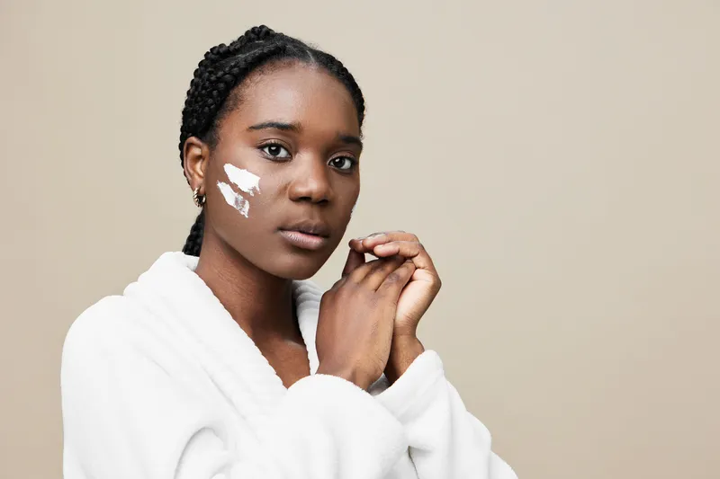 Young Black woman in a white bathrobe applying skincare cream on her face, showcasing a natural beauty concept with a neutral background and soft lighting