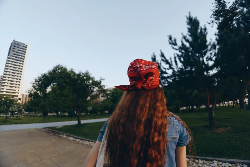 Lifestyle woman in modern boho-western denim street style with long red hair and red bandana walking outdoors, film color, urban park background, casual summer vibe