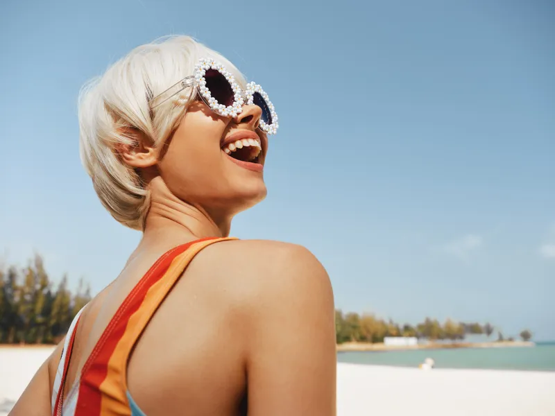 Joyful woman on a sunny beach wearing large sunglasses, bright colors, and a playful smile, capturing carefree summer style and vacation mood.