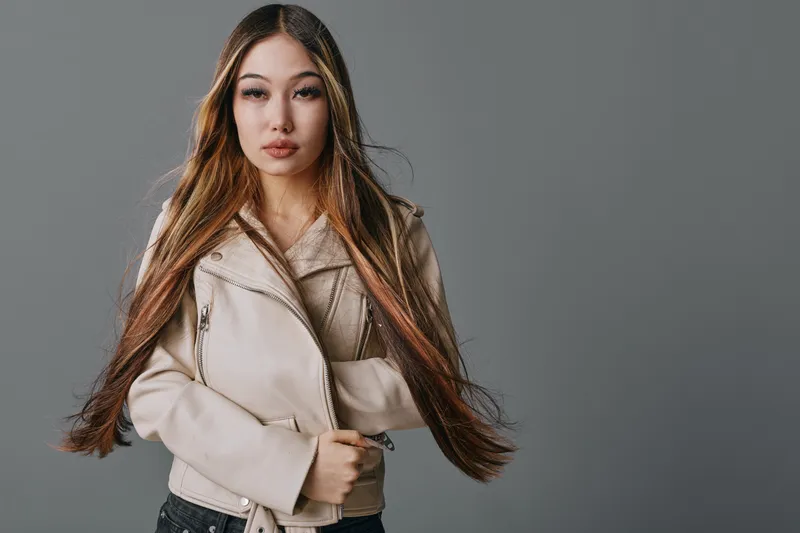 Young woman with long straight hair wearing a beige leather jacket stands against a neutral gray background with a confident expression and loose posture.