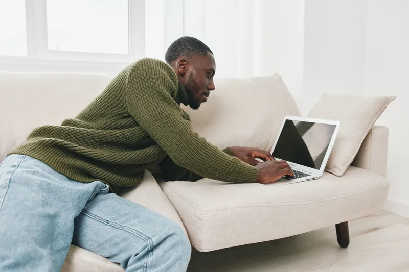 Young Black man working on a laptop at home, wearing cozy green sweater and jeans, relaxed pose, bright indoor setting, modern decor and seasonal comfort