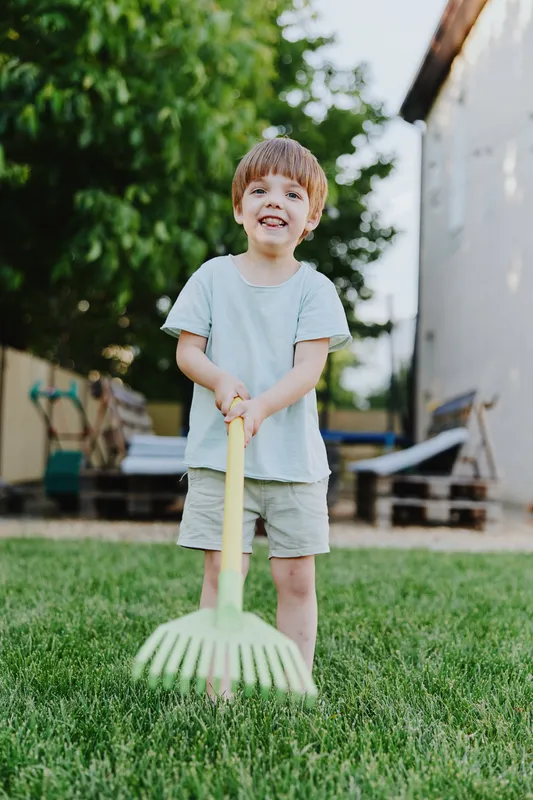Happy boy holding a garden rake, enjoying outdoor play in the sunny backyard surrounded by green grass and vibrant nature, perfect for family moments.