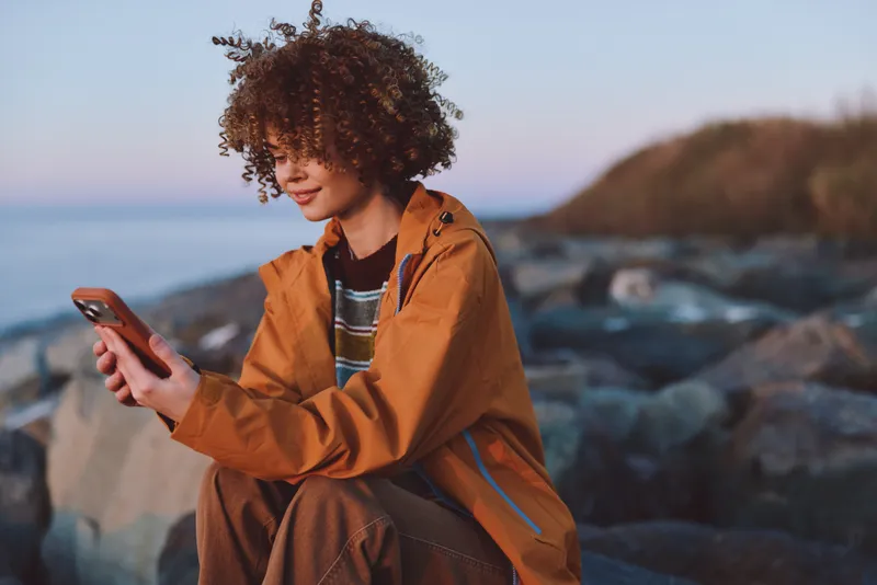 Woman with curly hair on a rocky coast, wearing an orange jacket, uses a smartphone and looks at the screen at sunset, outdoor lifestyle, communication, calm mood soft evening light