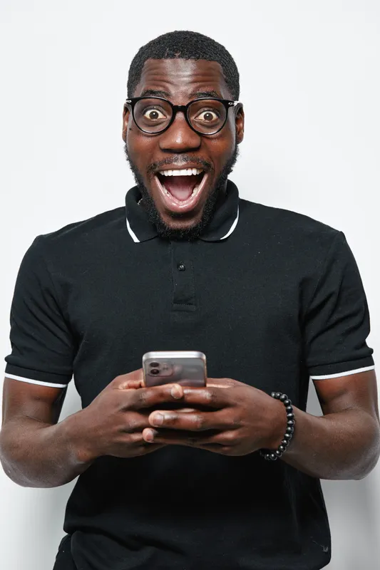 African American man with dark skin and glasses wearing black polo shirt holding smartphone with excited expression on white background in studio portrait.