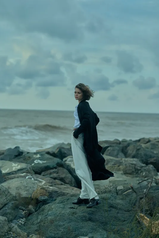 Coastal scene with wind sweeping through a long coat over a light dress, standing on rugged rocks beside the sea under a cloudy sky, evoking solitude, travel mood and natural contrast
