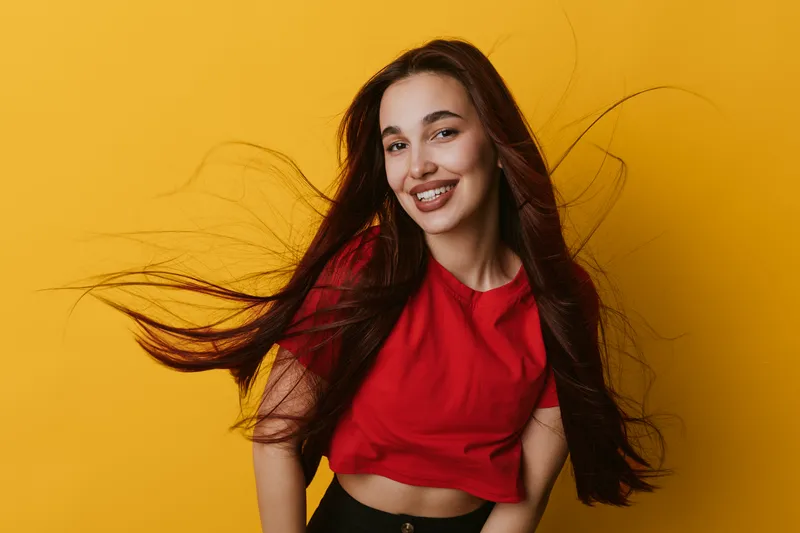 Young woman with long hair smiling and wearing red shirt on yellow background. Portrait of happy female with flowing hair and casual style in studio with vibrant colors.