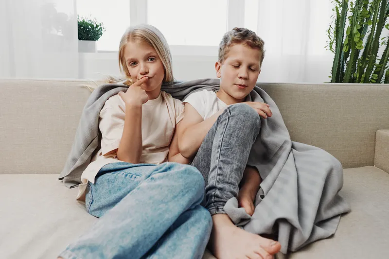 Two children sitting on sofa wrapped in blanket in cozy living room with plants and natural light. Relaxed mood of sibling bonding and casual home comfort captured indoors.