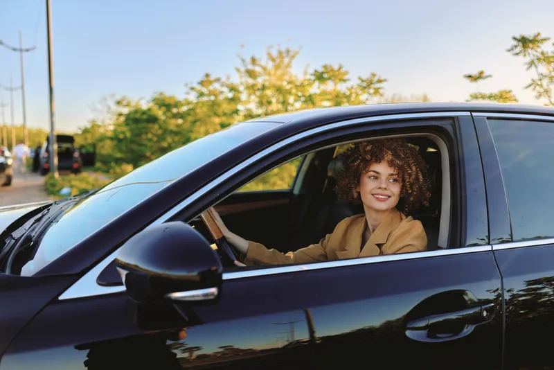 Woman driver sits in a sleek car during daytime, wearing a blazer, with retro film color grading, outdoor scenery, candid lifestyle moment, calm confident expression, stylish travel.