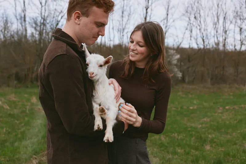 Cheerful couple outdoors with a small goat kid, affectionate interaction, casual clothing, rustic field setting, soft natural light, warm colors, candid lifestyle moment, joyful and relaxed