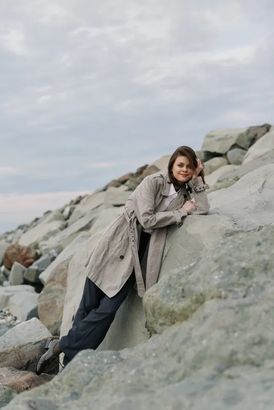 Young woman in warm coat leans on large rocks outdoors under cloudy sky. Casual fashion, nature, and relaxed mood with natural light on rocky landscape by the sea.