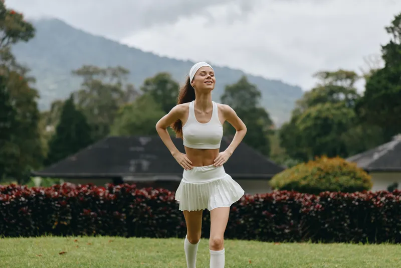 Young athletic woman in white sportswear jogging outdoors with a joyful expression, surrounded by lush greenery and mountains, emphasizing fitness and healthy lifestyle