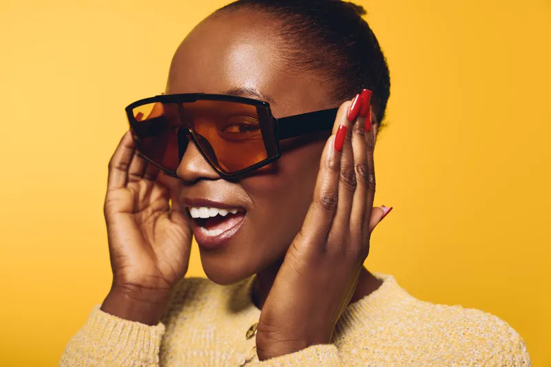 Smiling black woman with dark skin wearing oversized sunglasses and beige sweater poses on yellow background. Concept of fashion, style, beauty, and confident young African female model.