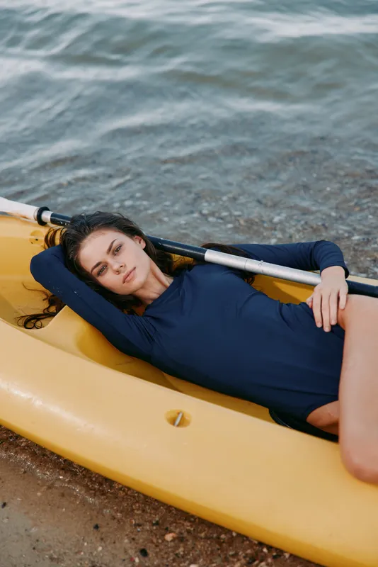 Female model reclining inside a yellow kayak along a quiet shoreline, wearing a dark blue long sleeve suit, sunlit water and sandy beach create a calm coastal scene.