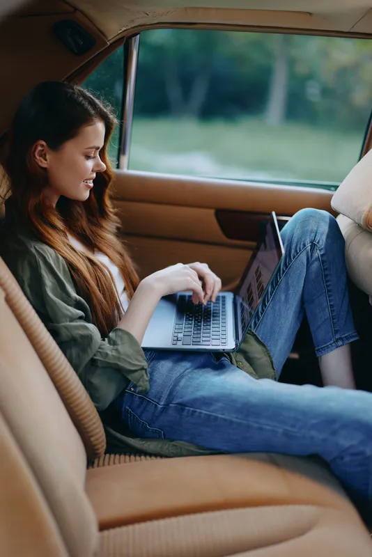 Young woman working on laptop in a car, dressed in casual outfit, conveying focus and creativity, with a green nature background.