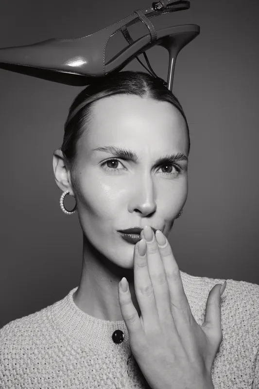 Caucasian woman with fair skin and dark hair holds a red high heel shoe on her head, covering mouth with hand in stylish studio portrait. Concept for fashion and elegance.