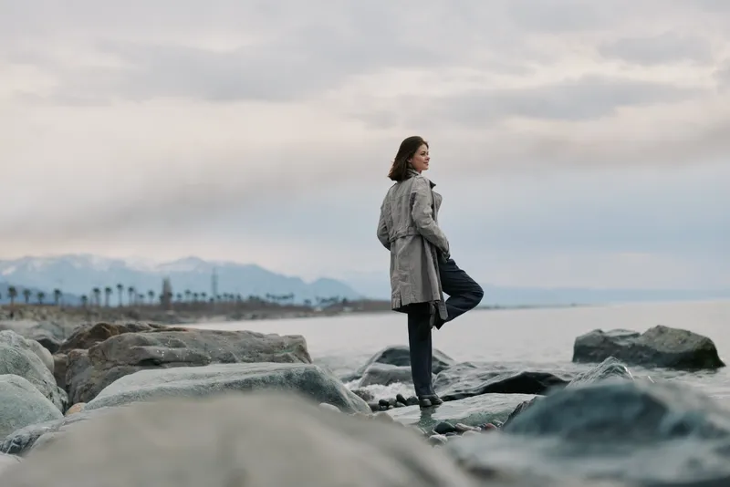 A woman in a grey coat stands on rocks by the sea, looking thoughtfully at the horizon under a cloudy sky. Coastal landscape with mountains and calm water.