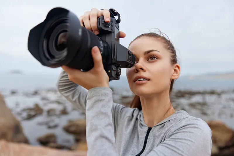 Young female photographer capturing nature scenes, wearing a gray hoodie, focused expression, outdoor setting with rocky background, suitable for lifestyle and photography themes