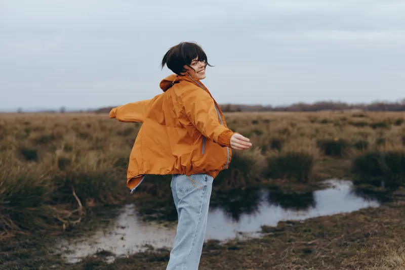 Woman lifestyle outdoors on a cloudy day, orange jacket and jeans, walking along a muddy path near a small river, hair blown by wind, arms adjusting the coat, countryside field