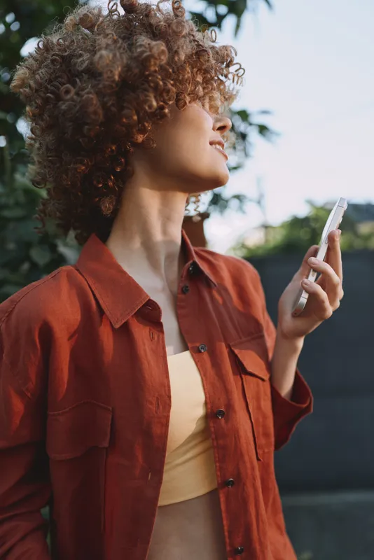A stylish woman with curly hair checks her smartphone outdoors, wearing a red shirt and yellow top, capturing a casual lifestyle moment in daylight