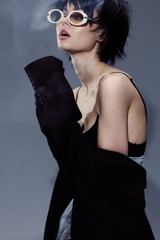 Stylish woman with short dark hair wearing oversized sunglasses and a black jacket, posing with an artistic and confident expression.