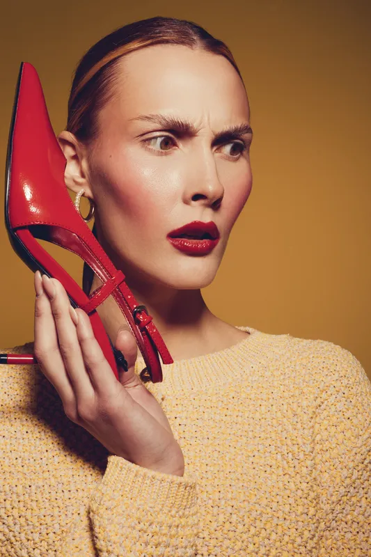 Woman with red lipstick holds red high heel shoe near her face, wearing yellow sweater on brown background. Concept of fashion, style, and unusual accessories.