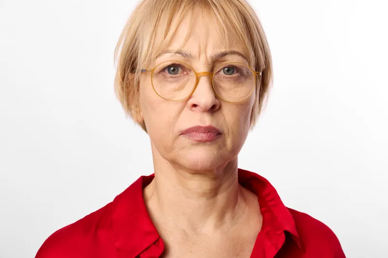 Senior woman portrait with short blond hair and round glasses wearing a red shirt against a white studio background neutral face expression calm confident mood