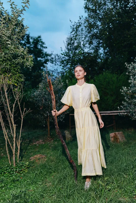 Woman in a pale dress stands in a lush garden holding a long branch, gazing forward with calm poise, daylight softens textures and creates a tranquil outdoor mood