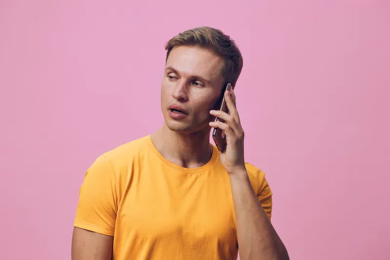 Young man wearing yellow T-shirt talking on mobile phone, looking serious and focused, standing against pink background in bright studio shot, casual fashion, lifestyle concept.