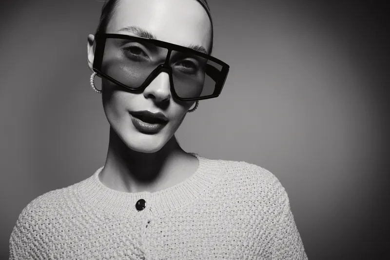 Elegant Asian woman with pale skin wearing large sunglasses and white knitted sweater, posing in black and white studio lighting.