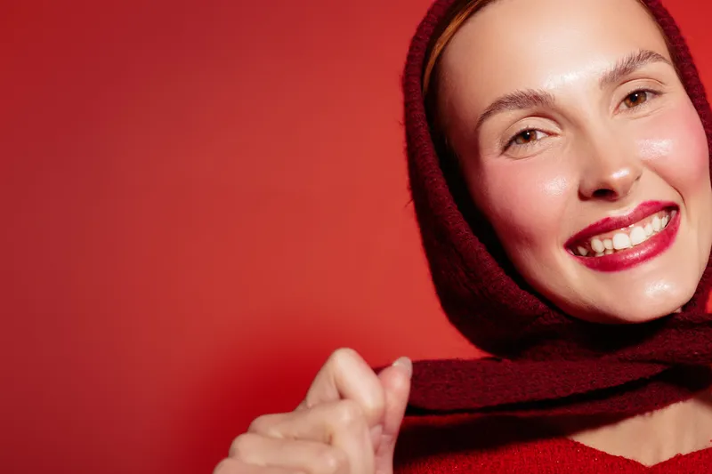 Smiling woman with red scarf on head poses against red background. Close-up portrait with natural makeup and bright lipstick. Concept of happiness, fashion, and beauty.