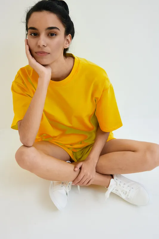 Young woman sitting cross-legged on white floor wearing bright yellow t-shirt and white sneakers, looking calm and thoughtful in casual indoor setting.