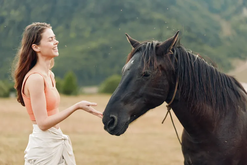 A cheerful woman stands in an open field, reaching out to a horse. She smiles warmly, creating a calm, friendly moment and a gentle connection with nature.
