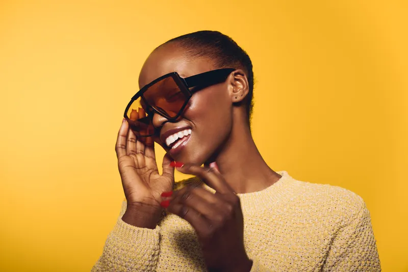 Smiling African woman with dark skin wearing large sunglasses and yellow sweater on yellow background. Portrait of happy black female model with short hair and positive mood in studio lighting.