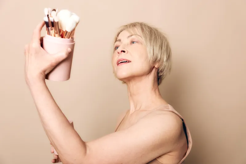 Mature woman with short blonde hair holds makeup brushes in a cup, looking upward with soft pink background. Concept of beauty, aging skin care, and self-confidence.