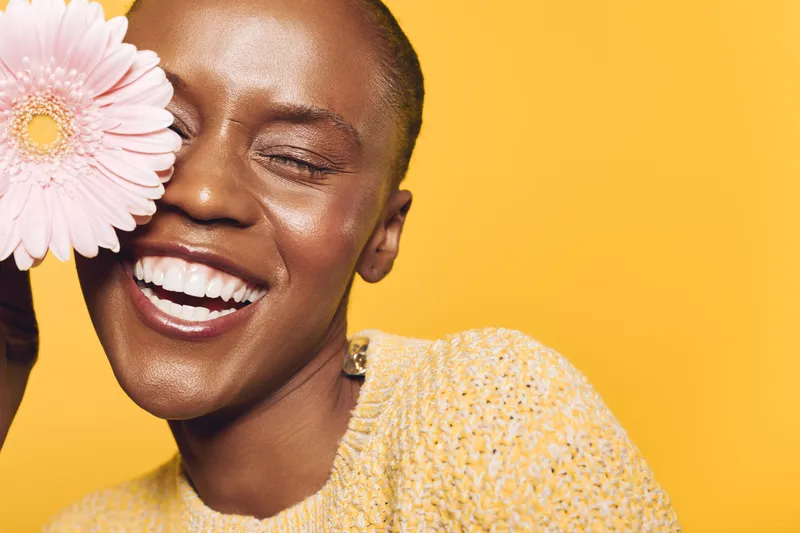 Smiling black woman with dark skin holds pink flower near face against yellow background. She wears yellow sweater and shows joy and natural beauty in bright studio lighting.