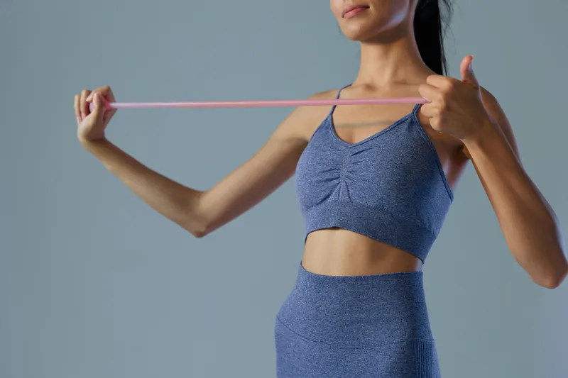 Woman sport fitness stretching with resistance band in activewear, focused exercise, toned arms, fitness training against neutral gray background.