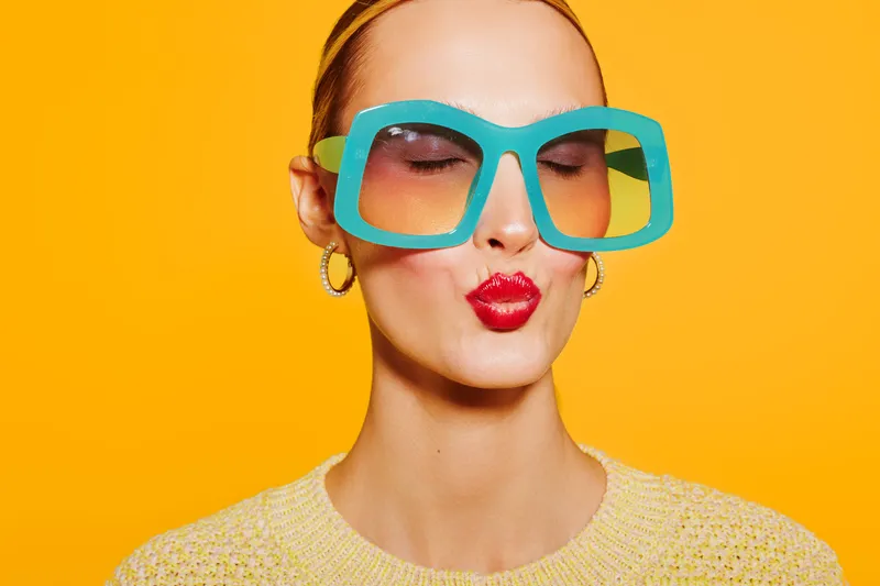 Young woman with large colorful sunglasses and red lipstick makes a kiss face on bright yellow background. Fashion portrait with trendy accessories and vibrant style concept.
