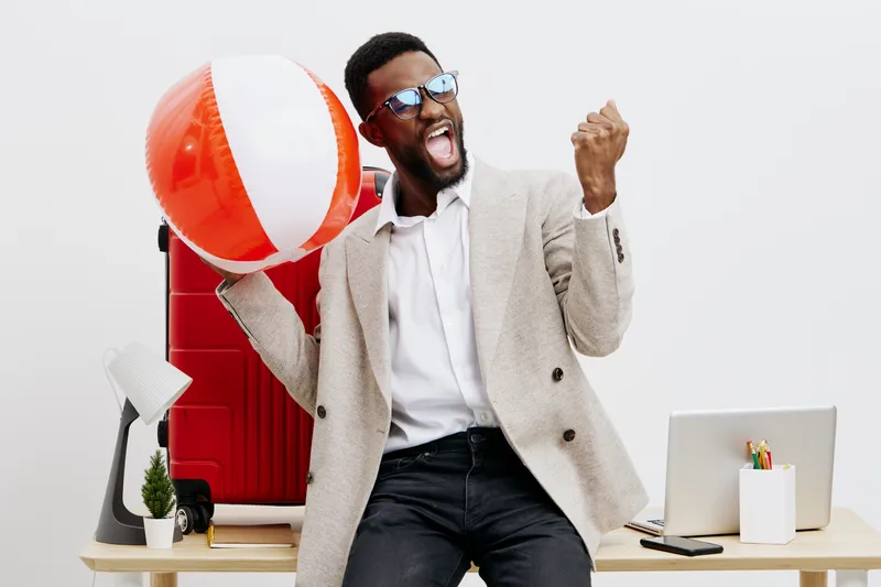 Excited cheerful young African-American man in smart casual clothing celebrating with a large beach ball in modern office environment, holding fist up in triumph, smiling, positive mood, lifestyle con