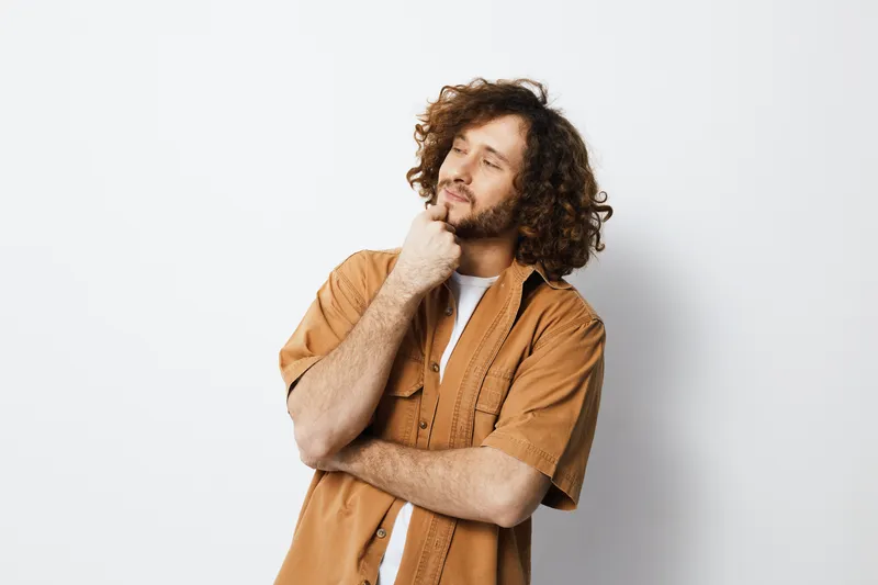 Pensive man with curly hair in a casual brown shirt, thoughtful pose against a minimalist white background, expressing contemplation and introspection