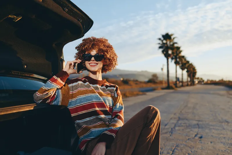 Woman lifestyle outdoors, smiling, sitting by car doorway, curly hair in warm sunset, relaxed posture, scenic road, palm trees, colorful knit sweater, golden hour breeze, travel freedom mood