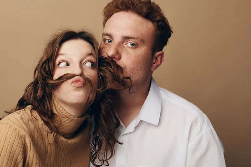 Playful young couple with red hair and freckles posing against beige background. Woman makes funny face with hair mustache while man looks at camera. Concept of fun, love, and relationships.