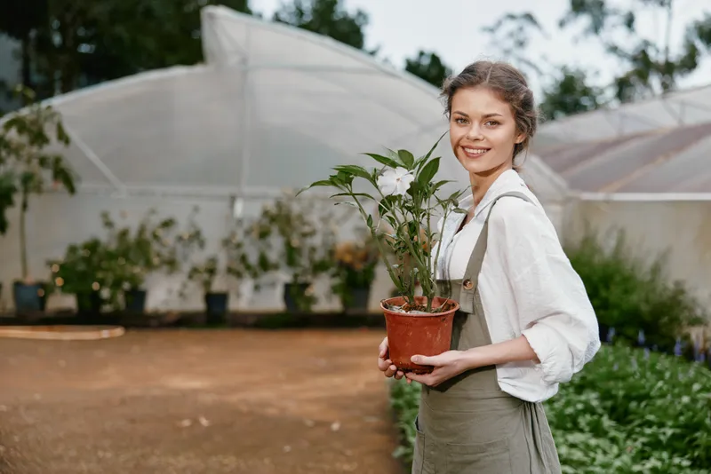 Young woman in casual outfit holding a potted plant in a greenhouse, showcasing joy and love for gardening while surrounded by vibrant greenery and natural light