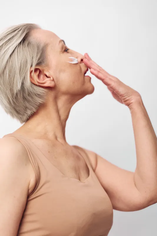 Mature woman with short grey hair applies moisturizing cream on her cheek, wearing beige tank top. Skincare concept, anti aging routine, self care and healthy skin care.