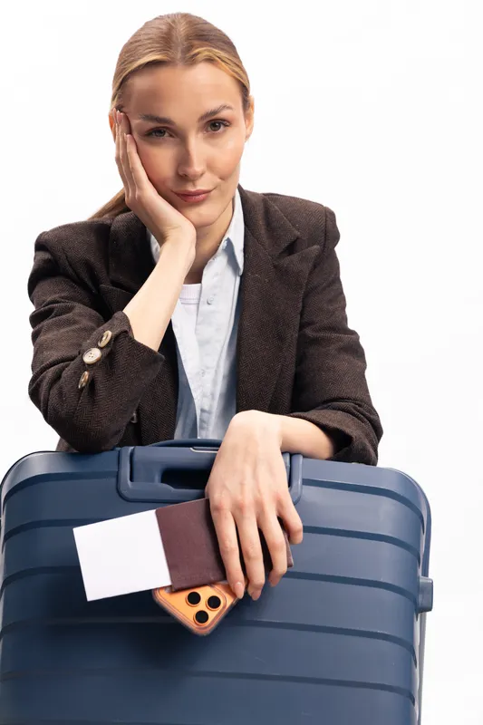 Young woman in dark blazer holding blue suitcase with passport and smartphone, looking at camera. Travel concept with business style, preparation, and journey planning on white background.